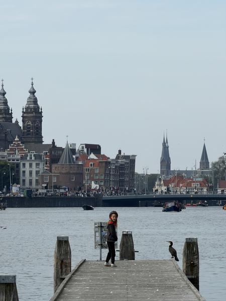 A young child stands on a wooden pier, looking towards the camera, with a cormorant perched nearby